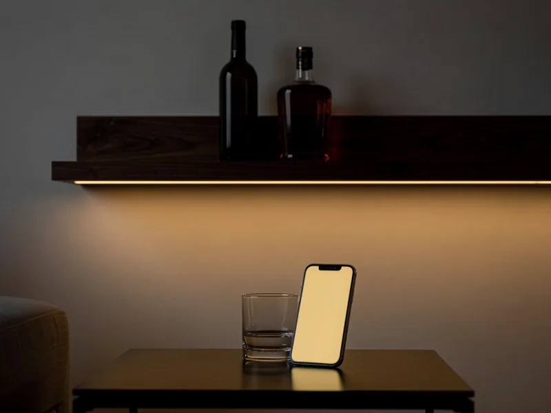 Weinflasche und Whiskyflasche ohne Etikett in einem modernen Regal, davor ein Smartphone mit warm leuchtendem, leerem Bildschirm